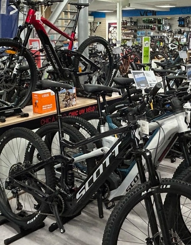 Magasin de vélos bien achalandé, avec VTT Moustache rouge et VTT électrique Scott noir. Présentoir "CHIC UN VÉLO".