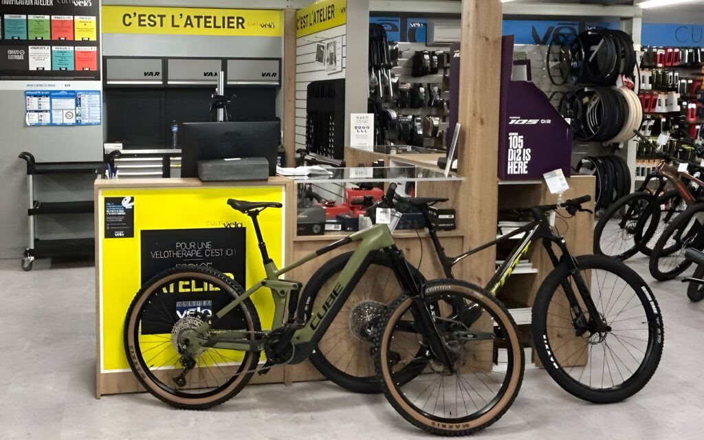 VTT Cube et Comptoir d'Atelier Vélo Professionnel Devant un comptoir d'atelier vélo, deux VTT Cube, dont un vert olive, exposés près d'affiches "C'EST L'ATELIER". Pneus Maxxis visibles.